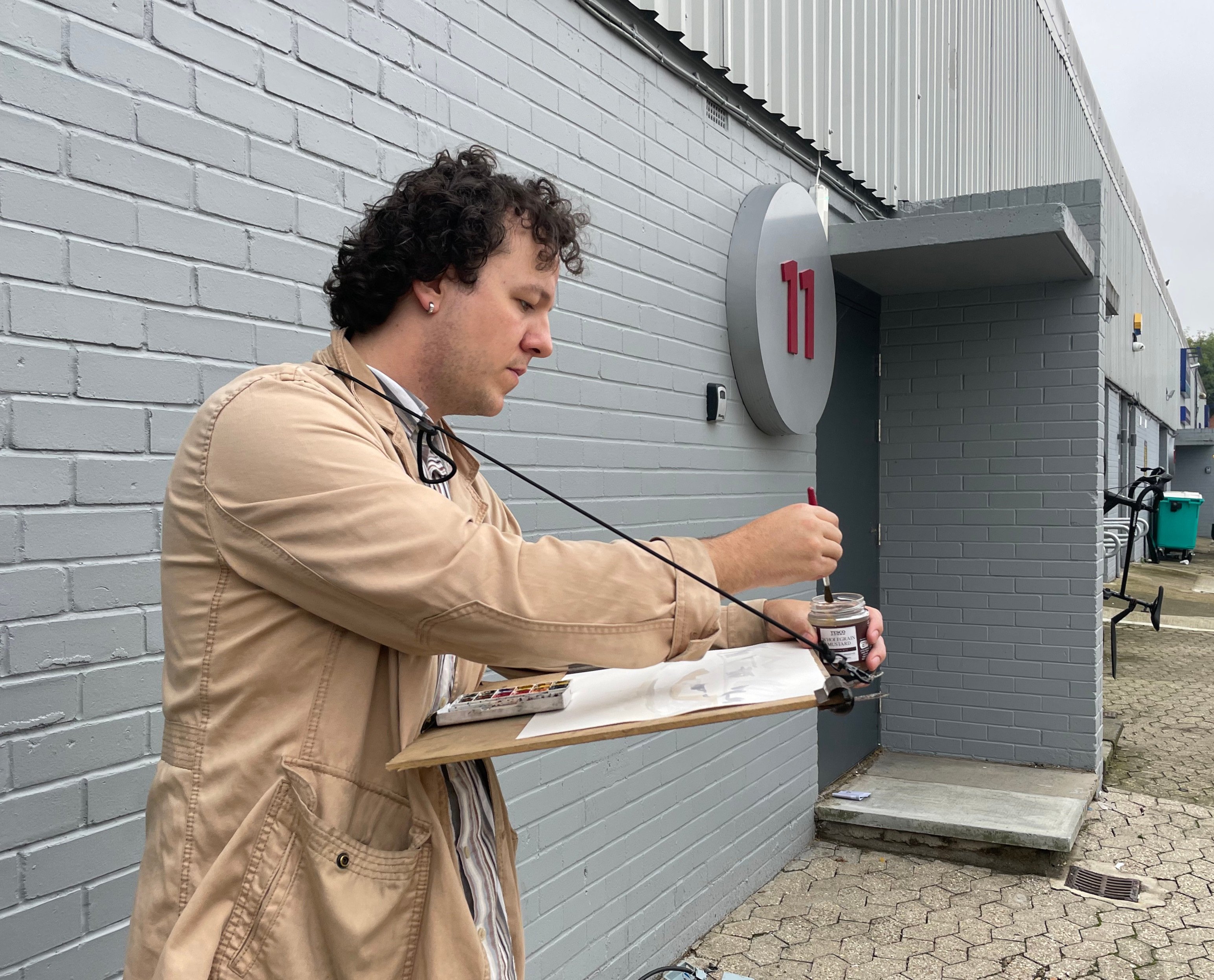 The artist Joshua Pell in a beige jacket sketches in front of a grey building