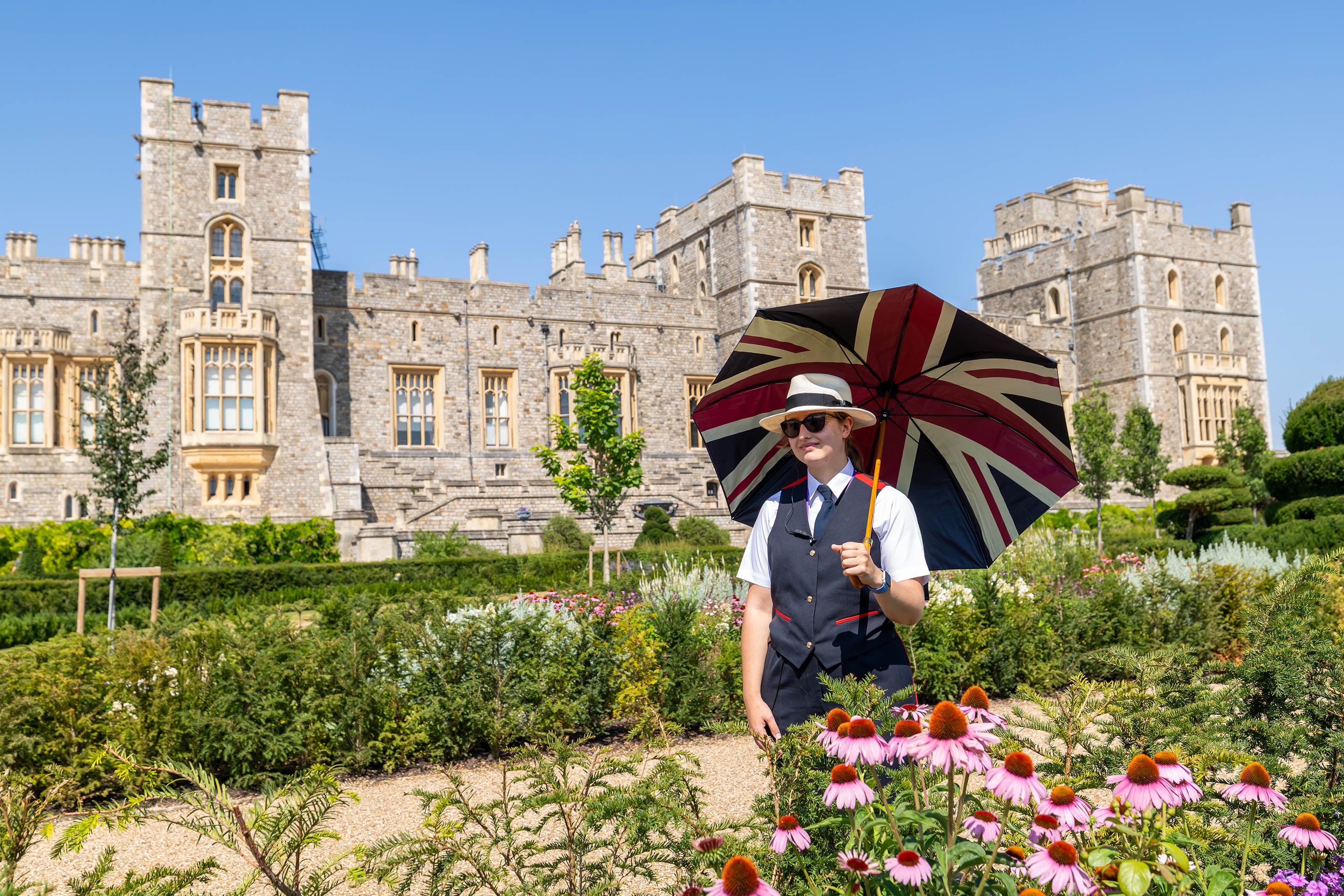 A warden in uniform holding a union jack umbrella in front of the east facade of Windsor Castle in the Venus Garden