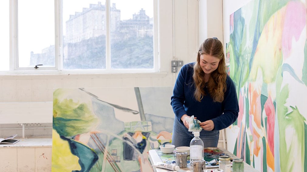 A woman with long hair and a navy jumper stands in an artist's studio surrounded by paint and colourful paintings