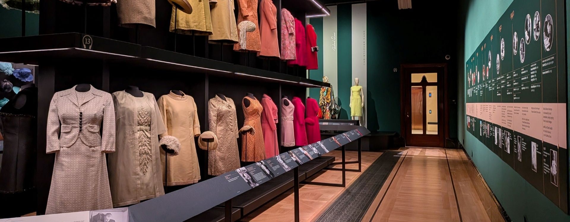 A green walled gallery with a selection of colourful dresses displayed on the left-hand side.