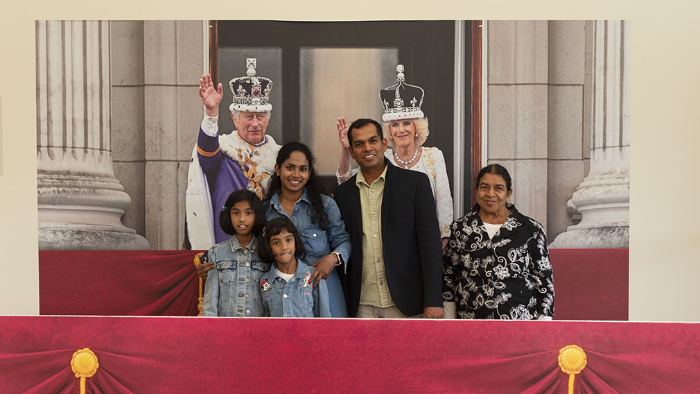 Adults and children are smiling standing in front of a picture of The King and Queen on the balcony of Buckingham Palace.