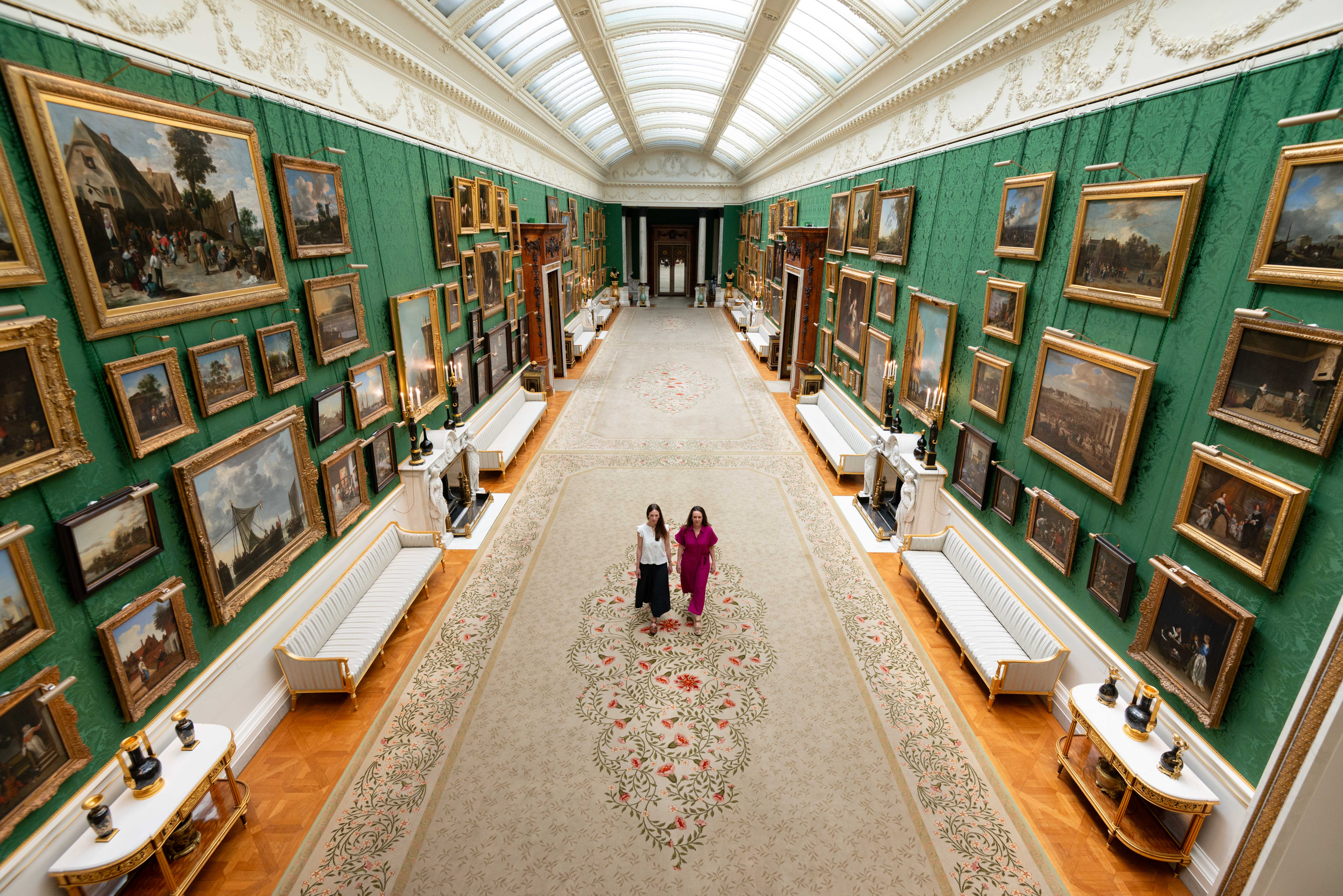 The newly rehung Picture Gallery at Buckingham Palace, a long room with green walls hung with lots of paintings and two people walking down the centre