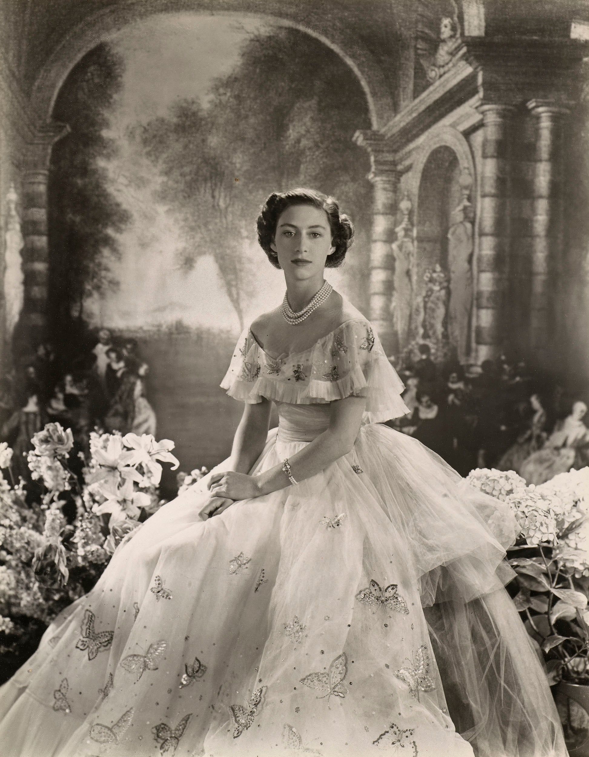 Princess Margaret with flowers posed against a painted backdrop (RCIN 2315232) Woman seated in white dress next to flowers.