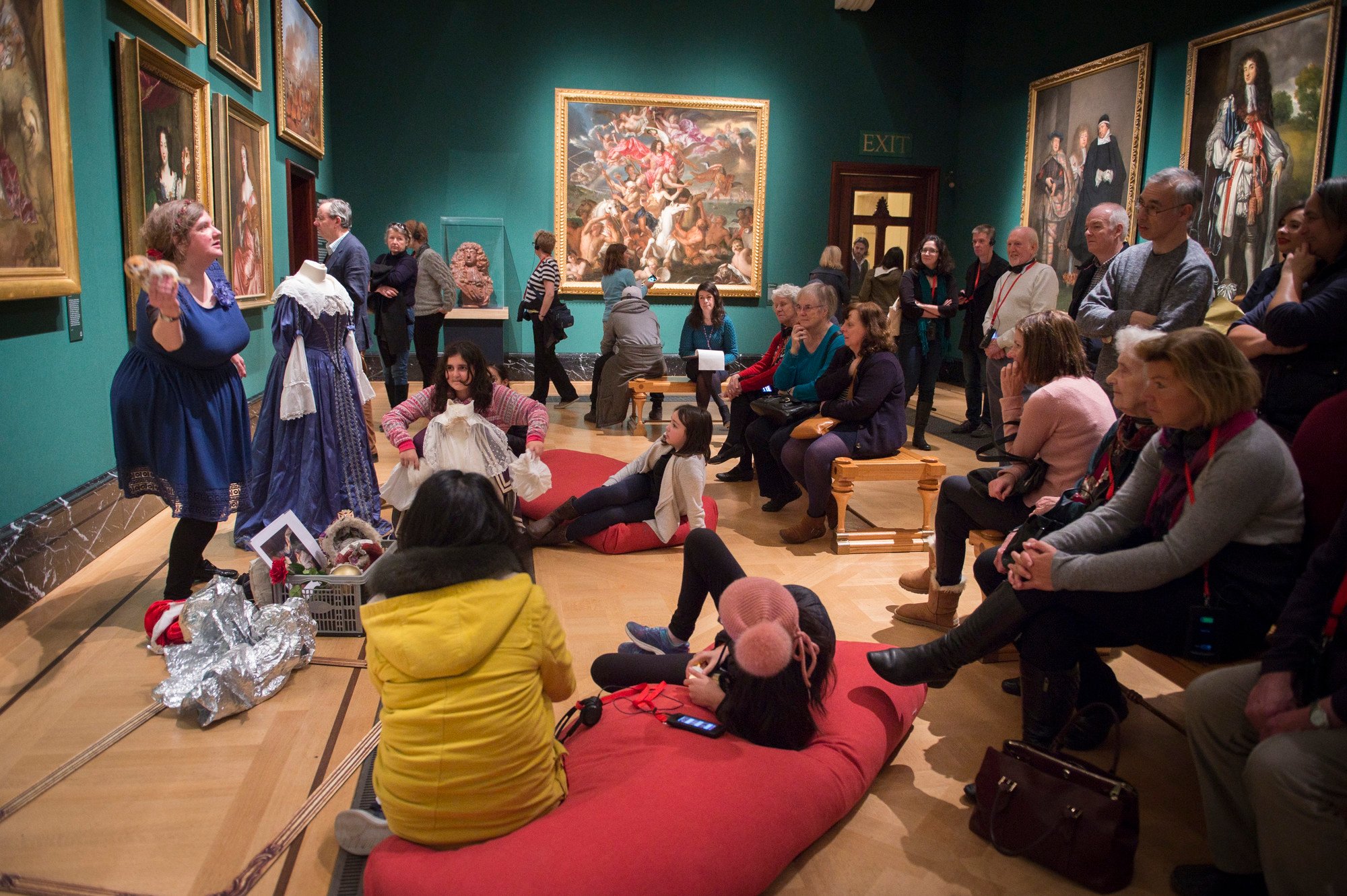 A woman is telling a story to an audience of adults and children in a museum gallery