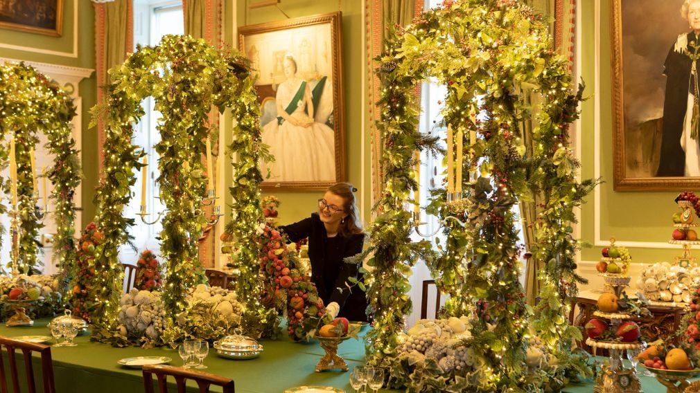 A person decorating a table with Christmas foliage and fruit. Paintings hang on the walls.
