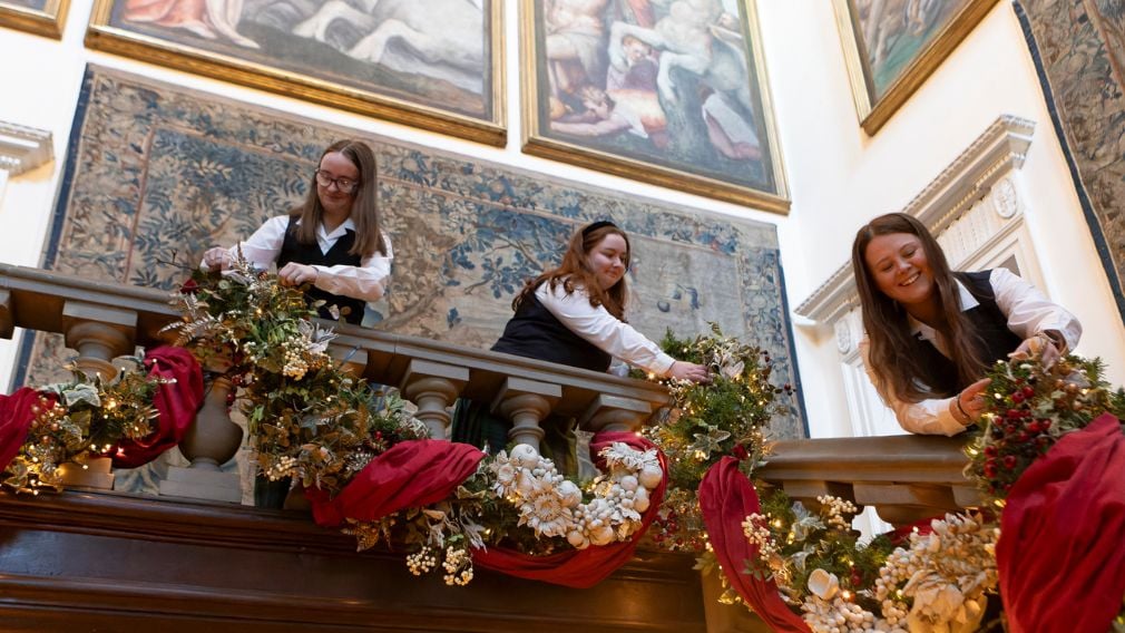 Three people adding Christmas decorations to a staircase. Tapestries on the walls behind.