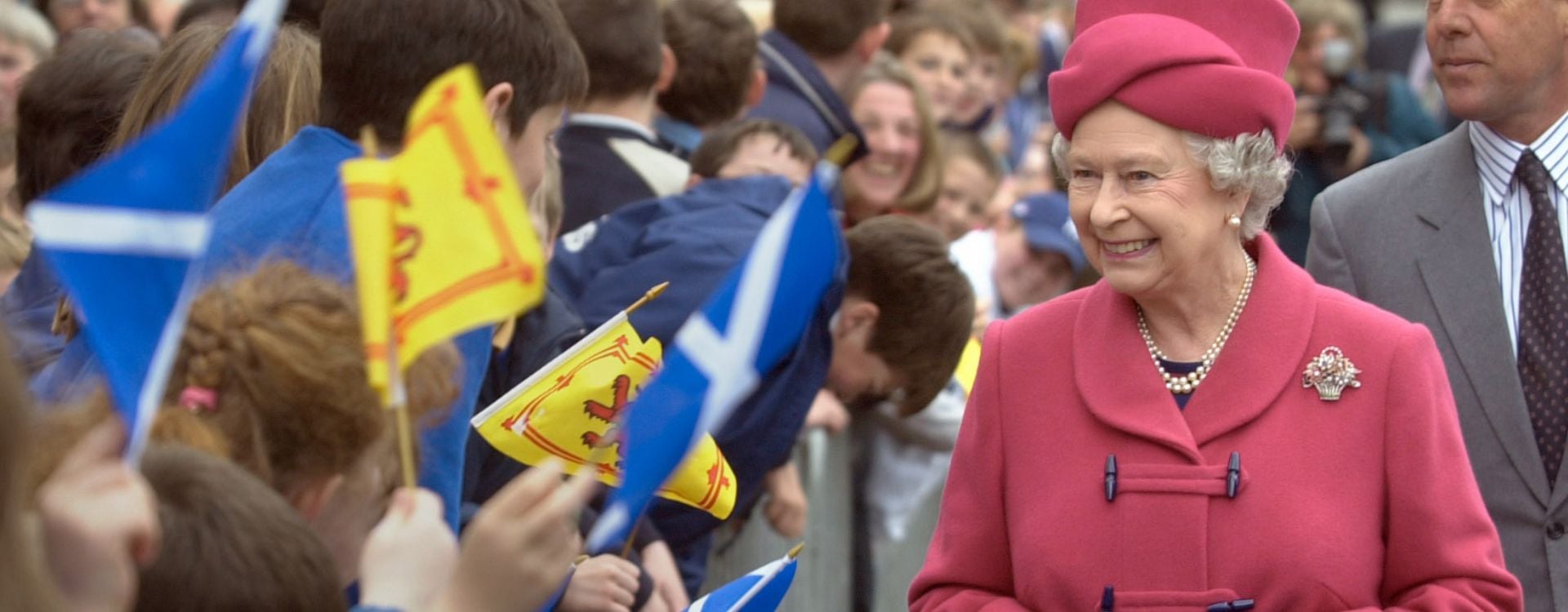Queen Elizabeth II in Scotland Queen Elizabeth II wearing a pink outfit walking through crowds waving Scottish flags.