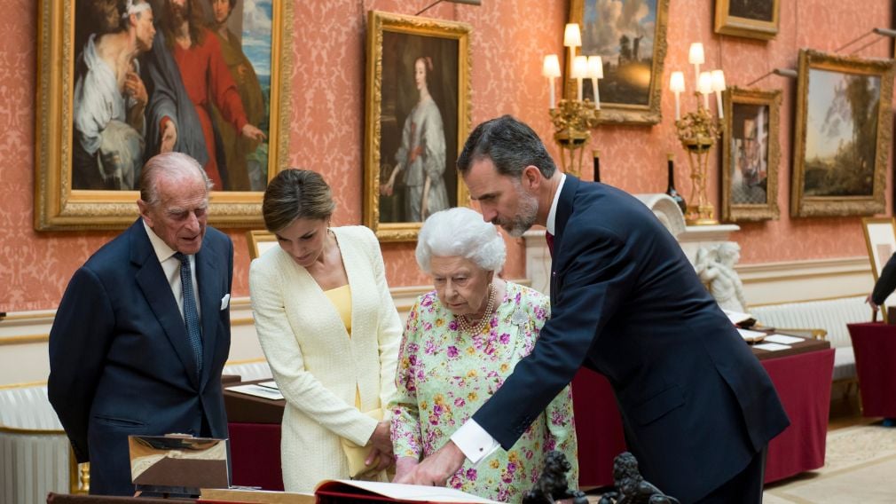 Queen Elizabeth II, Prince Philip, Duke of Edinburgh, Felipe VI of Spain, and Queen Letizia of Spain viewing a display in the Picture Gallery at Buckingham Palace during the Spanish State Visit in 2017. Four people look at a book on a table. Behind them are walls hung with paintings.