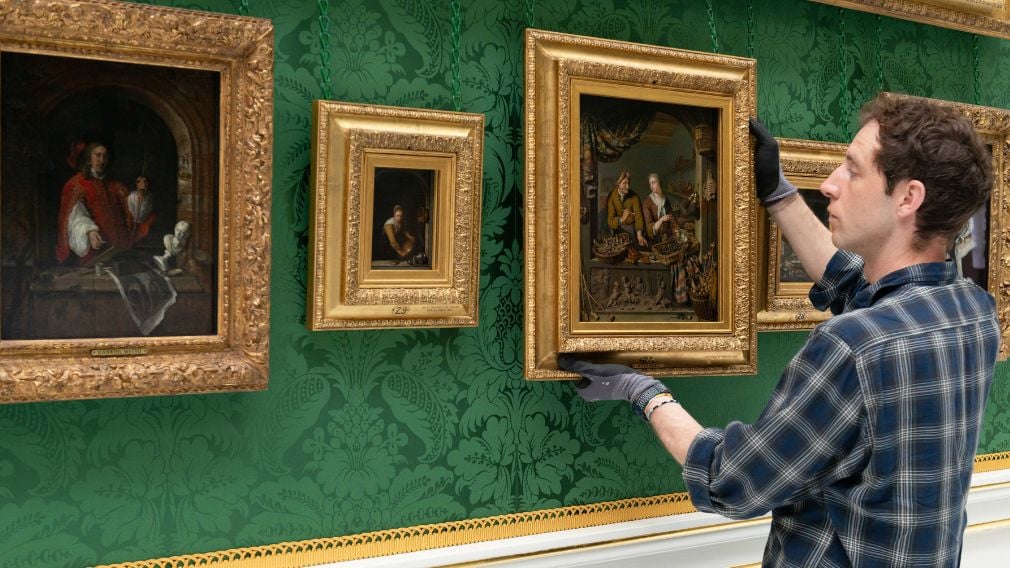 The smallest painting newly on display. A man hangs a painting on a green wall. Three more paintings hang on a wall.