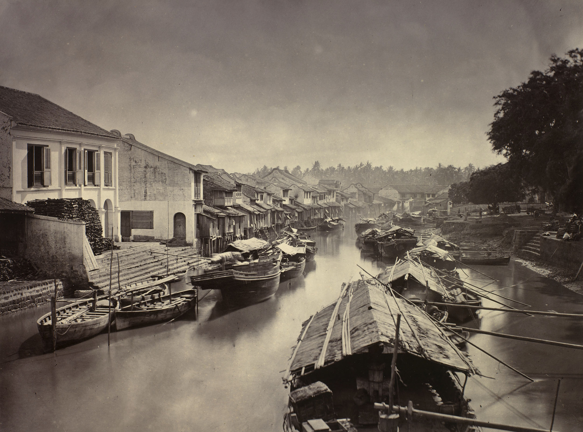Photograph of a view of the Melacca river, Malaysia. There are a large number of boats on the river, a selection with cabins and thatched roofs. On the left side of the river are a series of buildings, many of which are on stilts. Groups of people are sea