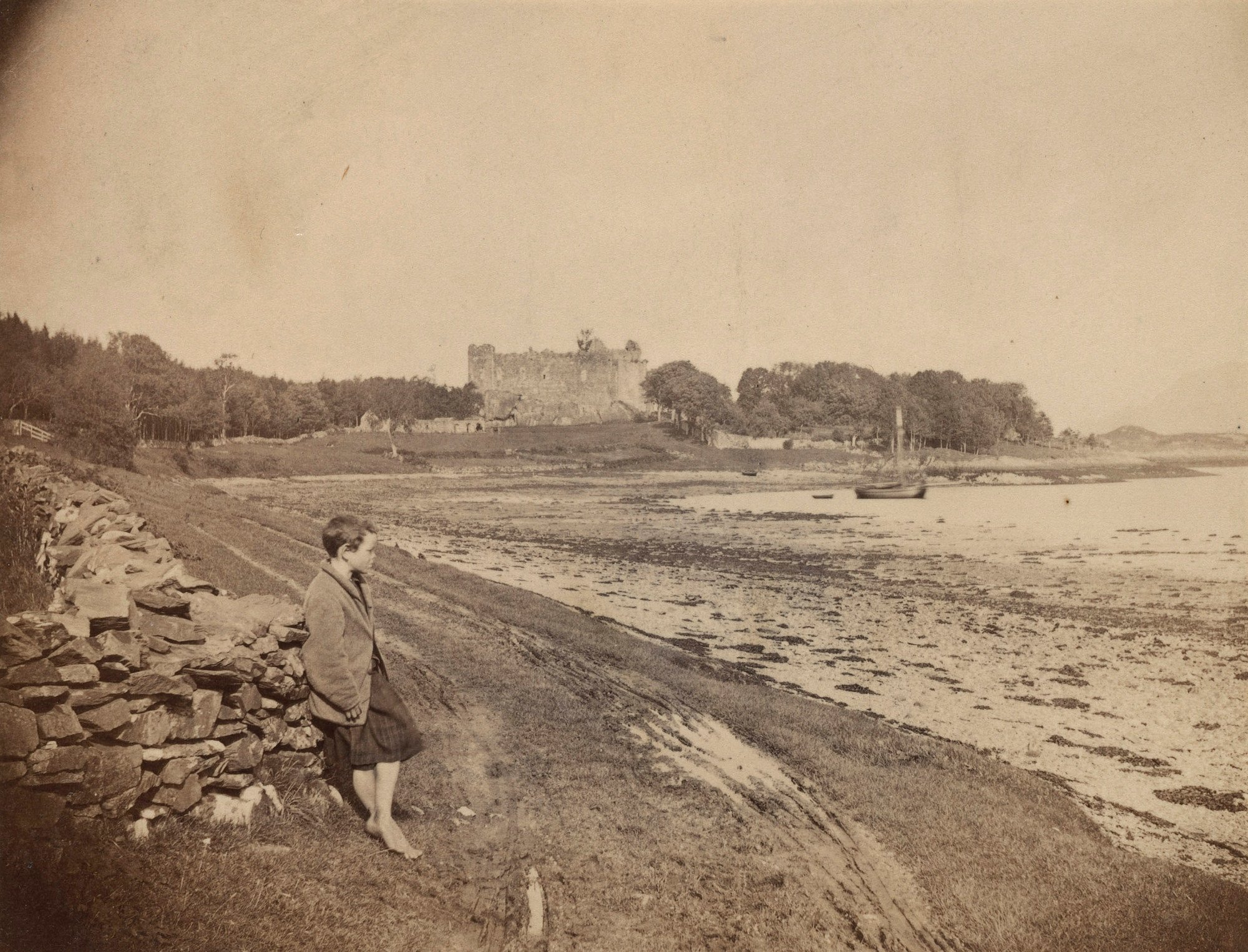 Photograph of a coastline with a young boy standing in right side profile as he leans against a&nbsp;low stone wall,&nbsp;looking out to&nbsp;sea. The boy wears three-quarter length trousers and is barefoot. In the background stands Dunstaffnage Castle on