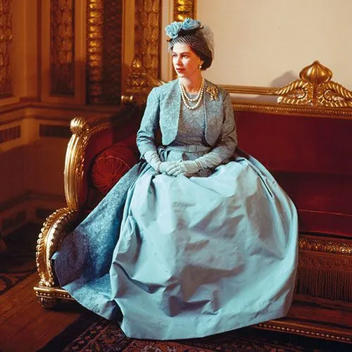 Queen Elizabeth II sitting down, wearing a blue gown and matching bolero jacket, and a hat featuring three large blue silk roses.