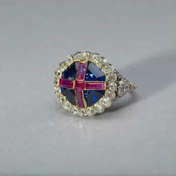 The ring comprises an octagonal step-cut sapphire, open-set in gold, overlaid with four oblong and one square rubies in gold strips forming a cross, within a border of twenty cushion-shaped brilliants in transparent silver collets. Brilliants decorate the