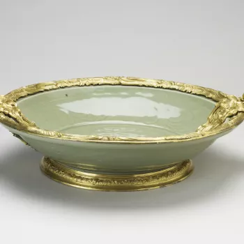 Chinese porcellanous stoneware dish, with carved decoration under a thick grey-green celadon glaze and French gilt-bronze mounts

Thickly potted, with narrow, spreading rim. Carved in the centre is a flower spray and round the sides a scrolling design, wi