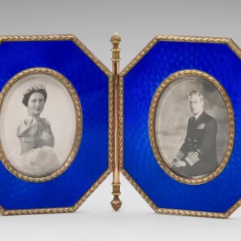 A Faberge double photograph frame, the two frames of octagonal shape of blue enamel. The frames of gold are joined by a hinge ending at the top and bottom in an acorn finial. The edge of the frames and oval reserves with laurel leaf border. Frames contain