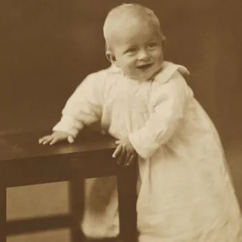 Photograph of the infant Prince Philip of Greece, later HRH The Duke of Edinburgh, aged approximately one year. He is pictured holding on to a wooden chair which he stands next to.