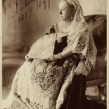 Photograph of Queen Victoria seated in three-quarters left position wearing a lace headdress with a coronet and holds a fan in her left hand. She is seated in a chair which stands upon a carpeted dais. Although this portrait of the Queen was used as the o