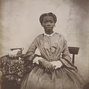 Photograph showing a three-quarters length portrait of Sally [Sarah] Bonetta Forbes, facing the viewer, seated and resting her right arm on a cloth-covered table. The portrait was captioned in the 19th century in ink 'Sally Bonetta Forbes' and dated 1856.