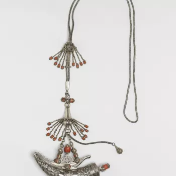 An Algerian powder horn of plain white metal with chased and pierced casing. The lid with a pierced and ornate coral finial. The clasp with three boss corals, the two swivels with five embossed corals each and two chains. The whole suspended from a heavy 
