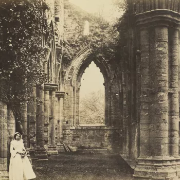Photograph of a young woman at Tintern Abbey. The woman stands in the foreground, on the left. She leans against an&nbsp;ivy covered pillar and looks towards the camera. The view is of an aisle that stretches to the background of the photograph with a win