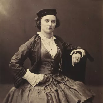 Photograph of a three-quarter length portrait of a woman, facing towards the viewer.&nbsp;She rests her left arm on the back of a chair and her right hand on her right hip. The woman wears a dark colour hat, light colour shirt, dark colour, fitted shirt, 