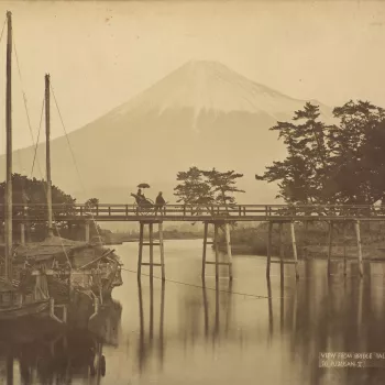 Photograph of a man pulling a jinrikisha (a Japanese type of rickshaw)&nbsp;with a&nbsp;passenger&nbsp;who holds up a parasol. The jinrikisha&nbsp;is depicted&nbsp;crossing the wooden Tagonoura Bridge, which is reflected in Fuji river. Two sailing barges 