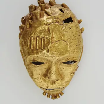 An Ashanti gold trophy head in the form of a hollow mask, with hammered facial features and pierced eyes, nostrils and mouth. Narrow strips of gold on the forehead and cheeks emulate scarification. Wirework coils, some missing, form hair curls and a beard