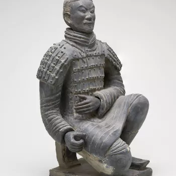 A kneeling Chinese warrior wearing body armour, his hair tied up in a bun and with a short moustache, sculpted in dark grey clay. The sculpture is a small-scale replica of the kneeling archer&nbsp;from the Terracotta Army of Emperor Qin Shi Huang (259-210