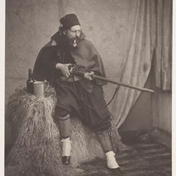 Photograph of Roger Fenton (1819-1869) dressed in traditional Zouave costume. He is leaning against a table which is covered by an animal fur, with his right leg raised off the ground. He is facing partly right and is holding a rifle with both hands. He i