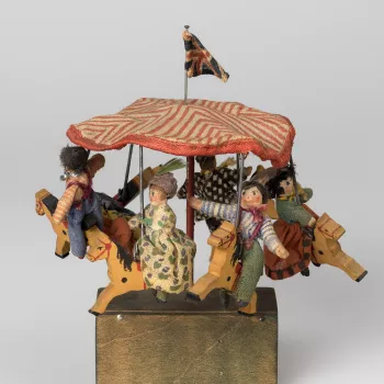Miniature toy merry-go-round with red&nbsp;and white striped cotton canopy with Union flag; from which are suspended six wooden painted horses each with wooden&nbsp;and textile rider. On green painted wooden base containing mechanism&nbsp;and turning hand