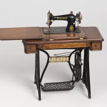 Miniature Singer sewing machine on integrated base of traditional form with hinged compartments (to lower machine), two small drawers and thread compartment at front.  Threaded for use.