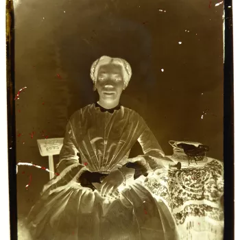 Glass plate negative&nbsp;of a three-quarter length portrait of Sarah [Sally] Bonetta Forbes (1843-80) seated, facing the camera front on. She poses with her hands held together in her lap. She wears a small hair covering, dark colour dress with lace coll