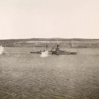 Photograph of HMS Queen Elizabeth&nbsp;under attack.&nbsp;The ship is at sea,&nbsp;the&nbsp;coast&nbsp;visible in the background. Two explosions cause large splashes&nbsp;in the water near the ship. The photograph was taken inside the Dardanelles on the s