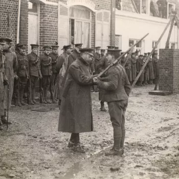 Photograph of King George V (1865-1936) in uniform and coat pinning a&nbsp;medal to the chest of a Sergeant from the 1st Division. The Sergeant holds a&nbsp;rifle over his left shoulder. The action takes place on a muddy street lined with buildings. A lin