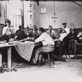 Photograph of Scots Guards at a tailors shop at the Chelsea Barracks. The soldiers are seated on stools around a large wooden table laid with sewing machines and various types of cloth. A selection of soldiers turn to face the camera and smile. A pile of 