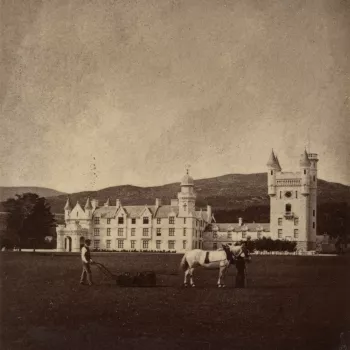 Photograph of two men in front of the new Balmoral Castle, Aberdeenshire.&nbsp;One leads a horse that pulls a lawnmower that the second man walks behind.