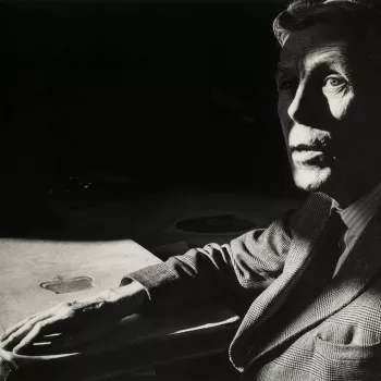 Framed photograph of a head and shoulder length portrait of&nbsp;Anthony Frederick Blunt (1907-83). He is seated&nbsp;at a desk&nbsp;gazing slightly upwards. A shadow falls across his face.&nbsp;A large book with a royal monogram is positioned on the desk