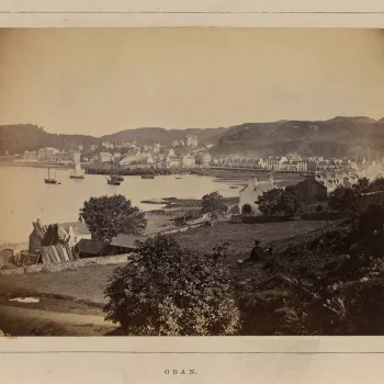 Photograph of the town of Oban with houses standing next to bay. Ships of varying sizes are on the water and an area of open grassland with a boy seated with his back to the viewer can be seen in the foreground.
Following the visit of Sir Walter Scott to 