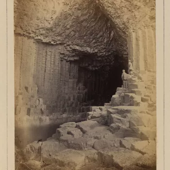 Photograph of the entrance to a cave formed of vertical basalt columns supporting a 'v' shaped roof. On the right of the entrance stand two men who face away from the viewer, looking into the cave. 
Photograph from a portfolio of 42 photographs taken by W
