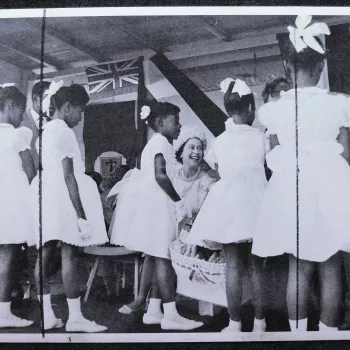 HM the Queen jokes with local children who stand with their backs to camera and wearing white dresses. The Queen attended the Harris Agricultural Show during her one day visit to Montserrat in the West Indies.