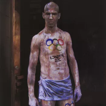 Colour photograph of a three-quarter length portrait&nbsp;of a man standing, naked apart from a blue coloured piece of cloth tied around his hips. He faces the camera front on, staring down the camera lens. He is covered is white and black paint and&nbsp;