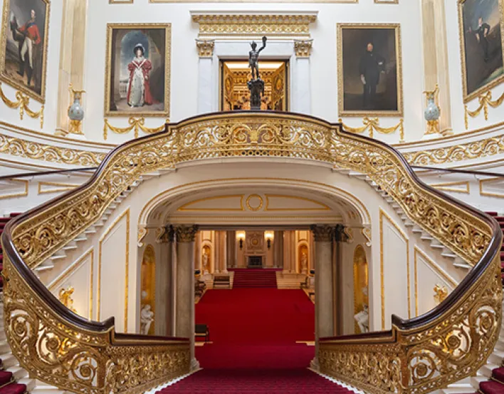 A double staircase with red carpet and gold banisters. The walls are lined with paintings.