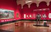 A gallery space with red walls, wooden floor, a wooden bench and exhibits including a suit of armour and paintings.