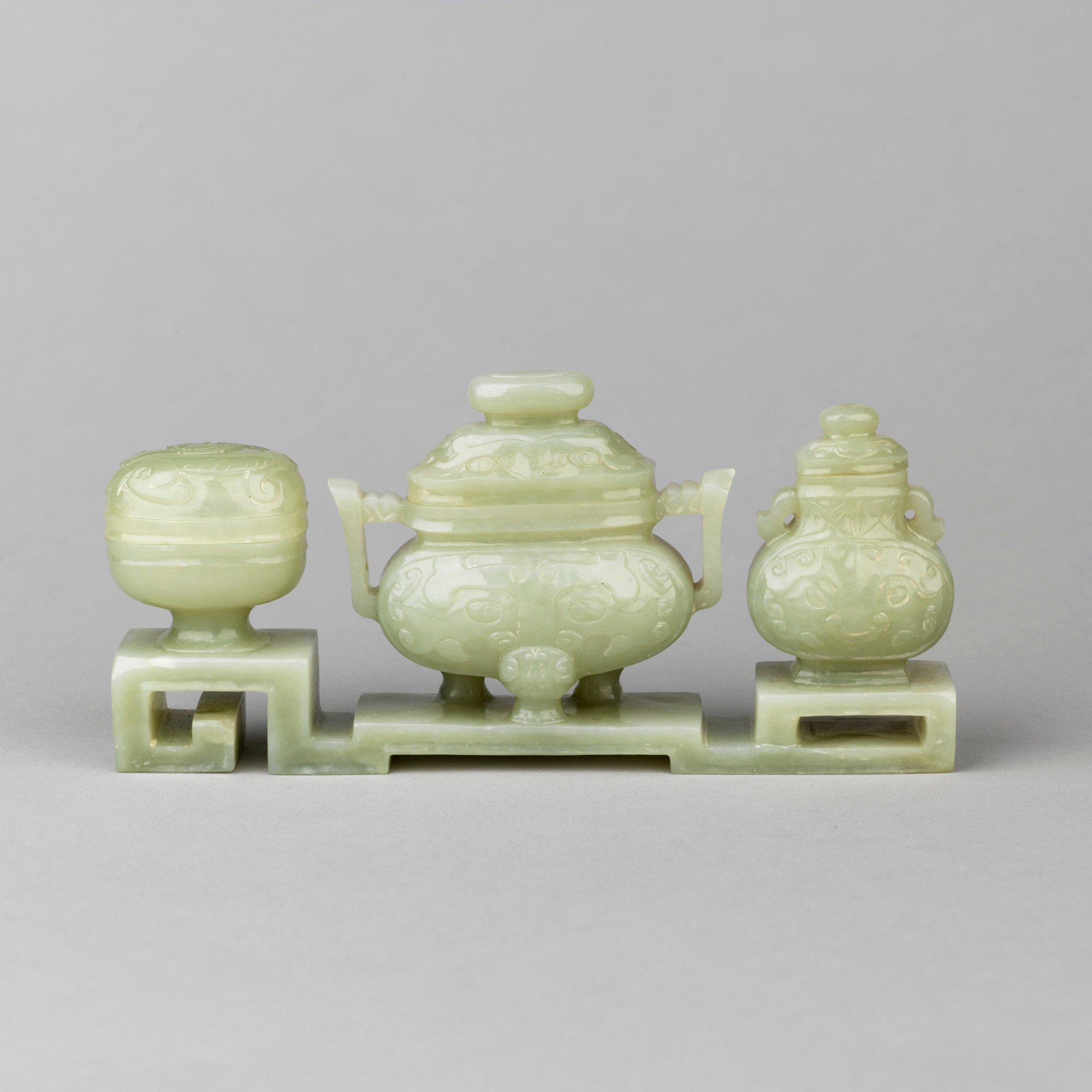 Three vases in archaic bronze forms carved on a stepped stand: in the centre, an incense burner, with upright handles and three animal-style feet, carved with a taotie, the domed cover with an oval button knob; to one side, a two-handled vase, also with t
