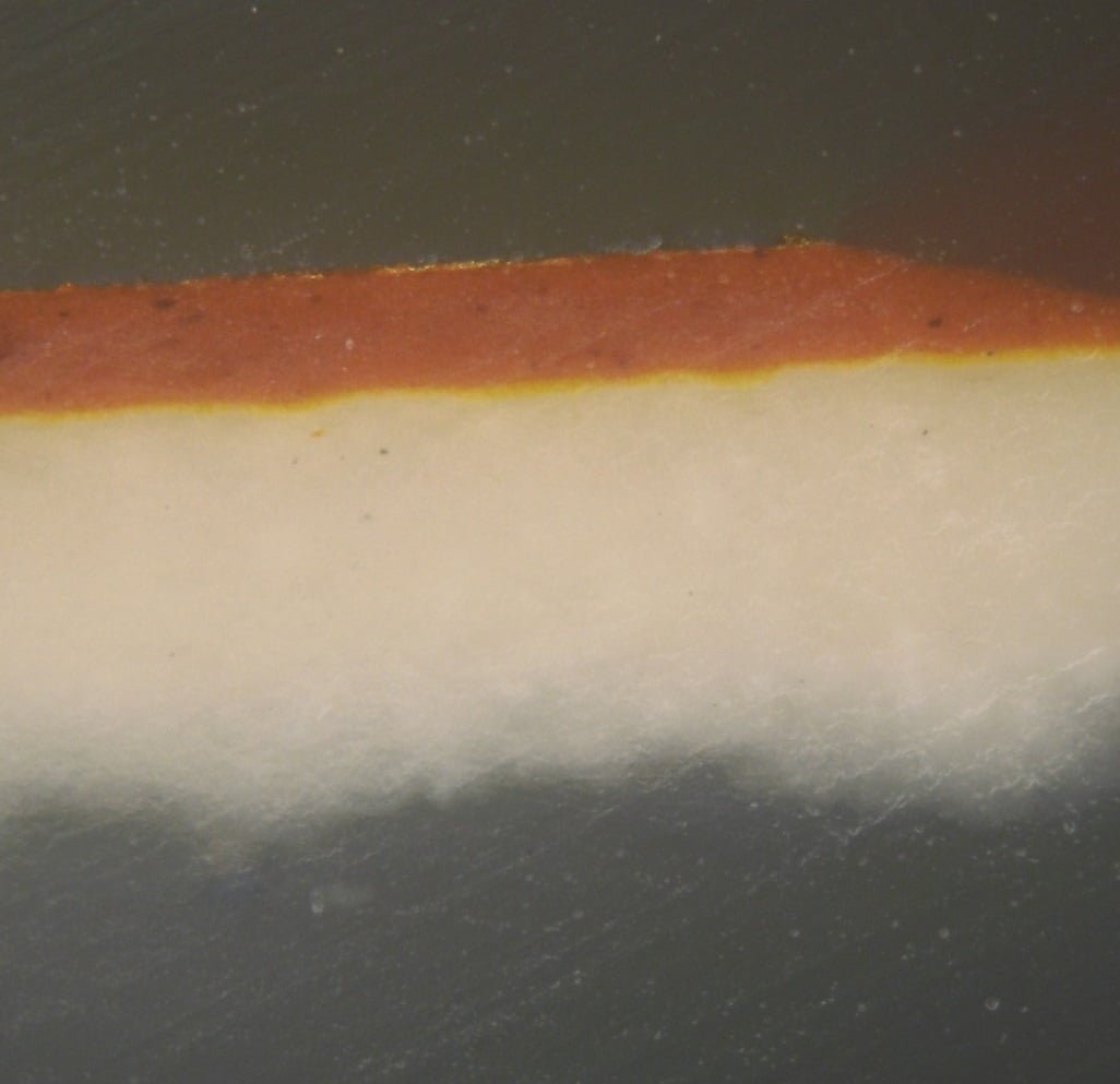 A cross-section of gilding, seen through a microscope. The component layers are clearly visible: gesso (in white), a thin layer of yellow ochre and glue, a red bole, and finally the very thin layer of gold leaf on top.